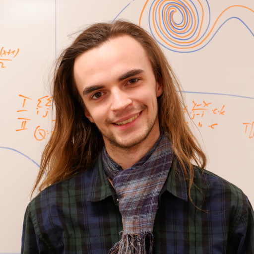 A smiling, white-skinned male-read person is facing the camera in front of a whiteboard filled with mathmatical symbols. They are wearing a scarf and shirt.